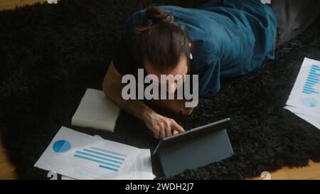 Foto di un giovane studente adulto con i capelli lunghi, che indossa cuffie wireless, sdraiato sul tappeto sul pavimento utilizzando una nota scritta da un computer tablet Foto Stock