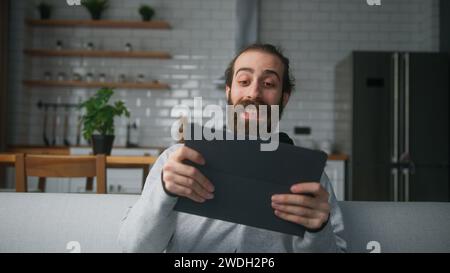 Un giovane uomo barbuto seduto sul divano a casa con lo sfondo della cucina, giocando a un eccitante concentrato impegnato in un interessante gioco mobil con un tablet Foto Stock