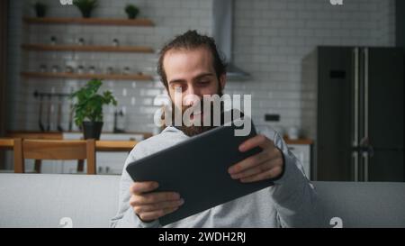 Giovane uomo barbuto con panino seduto sul divano a casa con sfondo in cucina, giocando emozionato concentrato coinvolto in un interessante gioco mobil Foto Stock