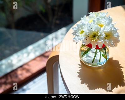 Vista dall'alto della bellezza dei fiori bianchi e gialli in acqua in vaso di vetro con luce e ombra a contrasto, decorazione su tavolo in legno rotondo vicino a gl Foto Stock