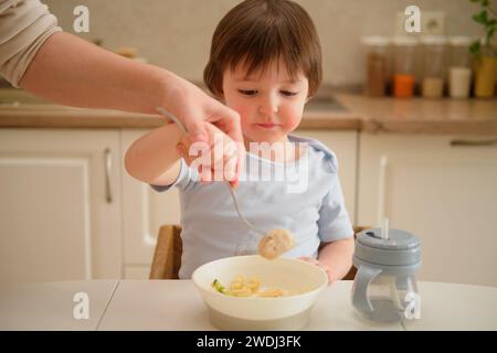 La madre insegna al bambino a mangiare con una forchetta in cucina. Mamma e figlio cucinano insieme in cucina. Bimbo di due anni a D. Foto Stock