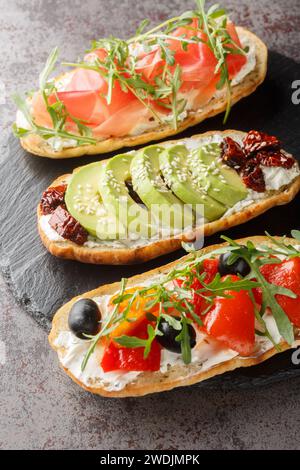 Bruschette assortite con rucola, prosciutto, pomodori secchi, avocado, formaggio spalmabile, primi piani di olive e peperoni arrostiti su una tavola di ardesia su t. Foto Stock