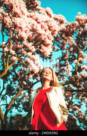 Bella e felice giovane donna che si gode una calda giornata di sole in giardino primaverile, indossa un abito rosso brillante e gli occhi chiusi Foto Stock