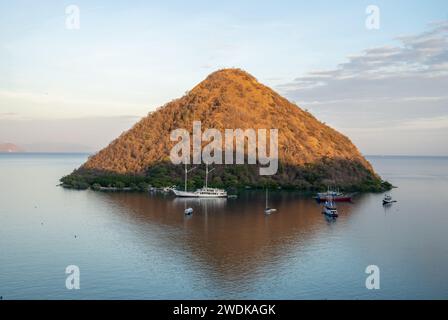 Splendore dell'ora d'oro: Una collina conica che sorge dalle calme acque di Labuan Bajo, Indonesia, con barche tradizionali ancorate nelle vicinanze Foto Stock