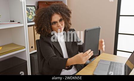 Una giovane donna ispanica con i capelli ricci che indossa occhiali e un blazer usa un tablet in un moderno ambiente da ufficio. Foto Stock