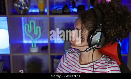 Una giovane donna ispanica con i capelli ricci che indossa le cuffie in una stanza da gioco buia di notte, che sembra concentrata. Foto Stock