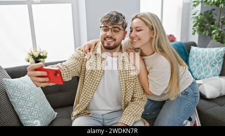 Una coppia amorevole scatta un selfie in un accogliente soggiorno dell'appartamento, mostrando affetto e interni moderni. Foto Stock