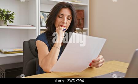 Una donna ispanica professionista di mezza età esamina attentamente un documento in un moderno ambiente di ufficio. Foto Stock