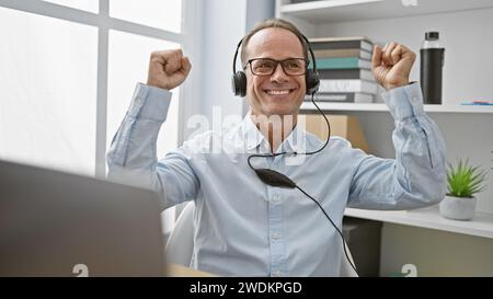 Allegro uomo d'affari di mezza età festeggia con un gesto vincente nel suo ufficio, ascoltando la musica accattivante sul suo computer portatile attraverso le cuffie Foto Stock