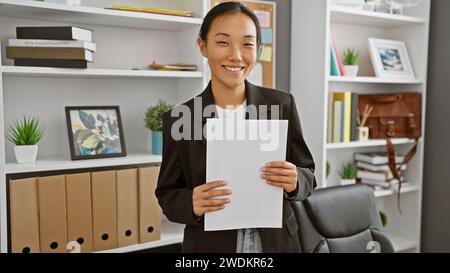 Una donna cinese professionista sorridente, che tiene i documenti in un moderno ambiente da ufficio con scaffalature e decorazioni. Foto Stock