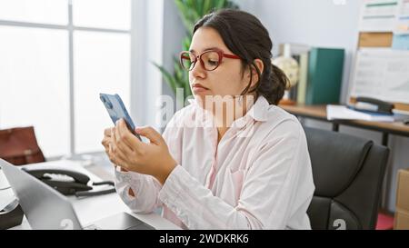 Una giovane donna ispanica con una camicia bianca e occhiali rossi usa uno smartphone in un ufficio con un laptop e uno sfondo della finestra Foto Stock