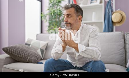 Bellissimo giovane ispanico, dai capelli grigi, seduto sul divano con un'espressione seria, profondo pensiero nel suo appartamento interno Foto Stock