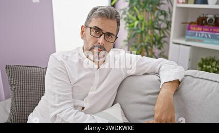 Un giovane ispanico con la barba dai capelli grigi, che indossa gli occhiali, rivela un'espressione seria mentre è seduto sul divano del soggiorno, in casa. Foto Stock