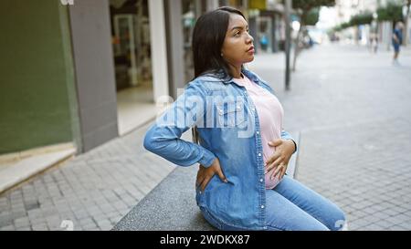 Una giovane donna incinta stanca, toccando la pancia, siede in contemplazione su una panchina urbana, rilassata, bilanciando l'emozione della maternità in attesa Foto Stock