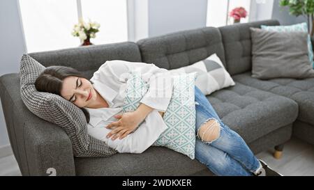 Una giovane donna con un'espressione dolorosa giace su un divano, stringendo lo stomaco in un soggiorno ben arredato. Foto Stock