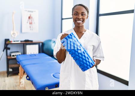 Donna afro-americana fisioterapista sorridente sicuro tenendo rullo di schiuma alla clinica di riabilitazione Foto Stock