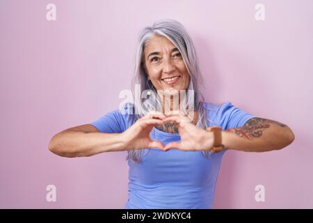 Donna di mezza età con tatuaggi in piedi su sfondo rosa sorridente innamorata che fa forma a simbolo del cuore con le mani. concetto romantico. Foto Stock