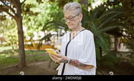 La donna anziana dai capelli grigi si concentra seriamente sul messaggio online, in mezzo al verde del parco illuminato dal sole, mostrando uno stile di vita maturo e esperto di tecnologia. Foto Stock
