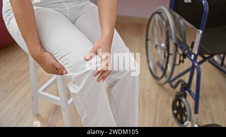 Una persona con pantaloni bianchi siede vicino a una sedia a rotelle in una clinica di riabilitazione, il che implica il recupero da lesioni o interventi chirurgici. Foto Stock