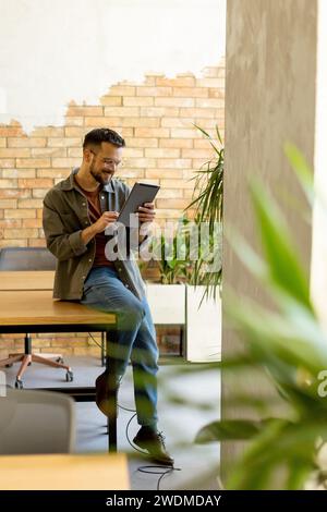 L'uomo allegro sta con sicurezza tenendo un tablet digitale in uno spazio ufficio contemporaneo con una parete in mattoni a vista, a simboleggiare una miscela di tecnologia moderna Foto Stock