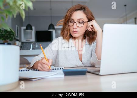 Bella donna di mezza età in occhiali che fa segni in documenti finanziari a casa. Immagine di concetto per piccole imprese, finanze domestiche, risparmio di denaro. Foto Stock