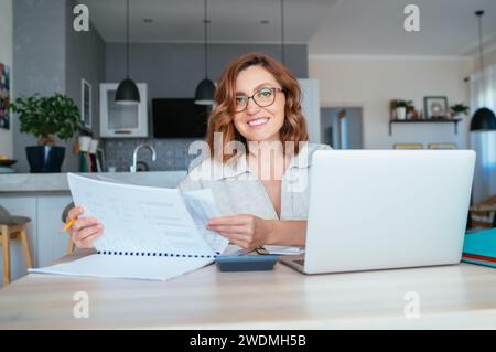 Bella donna di mezza età con gli occhiali che sorride alla macchina fotografica. Esegue rapporti finanziari trimestrali presso l'ufficio domestico utilizzando un moderno notebook. Piccole e medie imprese, Foto Stock