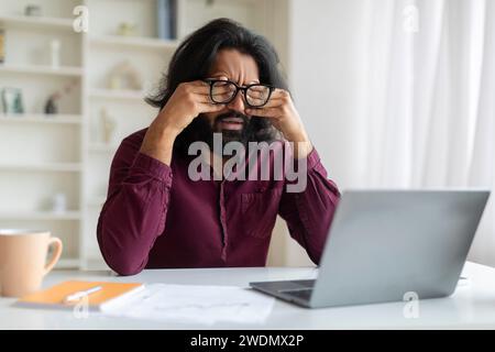 Uomo indiano stanco che si strofina gli occhi, affetto da affaticamento oculare durante il lavoro su un notebook Foto Stock