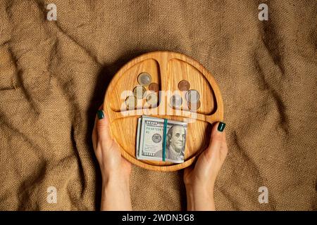 Le mani femminili tengono un piatto di legno con dollari e monete su uno sfondo di tessuto marrone, affari e finanza Foto Stock
