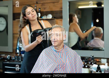 Il giovane parrucchiere professionale pettina e asciuga i capelli agli anziani nel barbiere Foto Stock
