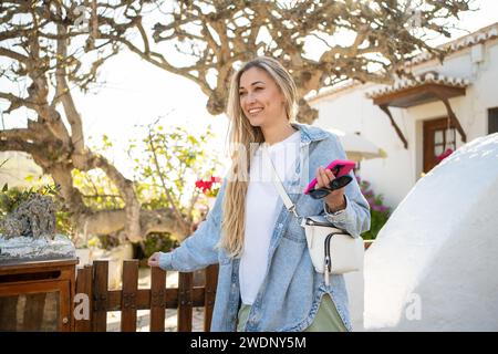 Giovane turista sorridente in piedi vicino alla casa bianca. Bella donna bionda con camicia in denim appoggiata su una recinzione di legno. Lady ha uno smartphone e occhiali da sole fuori dalla casa di campagna in Portogallo. Foto Stock