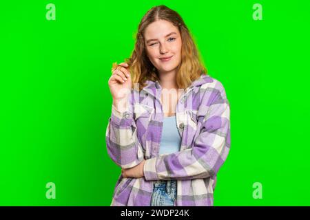 Sorridente allegra giovane donna caucasica che lampeggia gli occhi, guarda la macchina fotografica con sorriso, strizza l'occhio, flirta, esprime ottimismo, buone notizie. Affascinante ragazza rossa isolata su sfondo cromatico verde Foto Stock