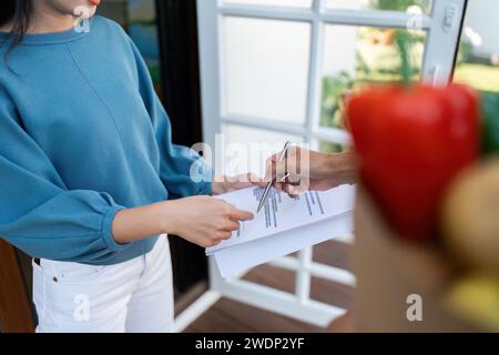 Asiatico consegna uomo che tiene la penna e maneggia il sacchetto di cibo, frutta e verdura dare alla cliente femminile di fronte alla casa Foto Stock