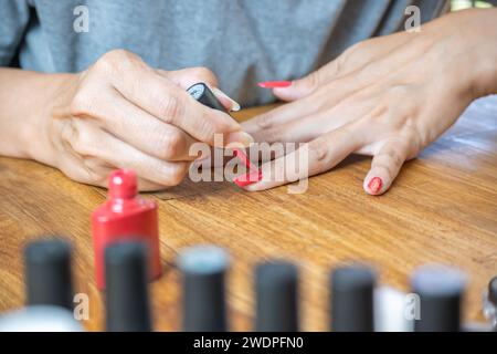 Mani di una giovane donna latina che dipinge le unghie con uno smalto rosso sul tavolo, circondata da accessori per la pittura. Foto Stock