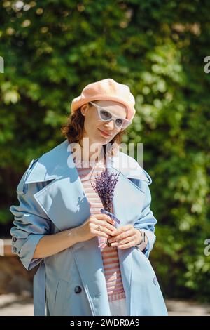 Donna alla moda con impermeabile blu e berretto di corallo pallido tiene in mano un bouquet di lavanda Foto Stock