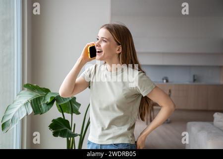 Simpatica donna sorridente in piedi vicino alla finestra, che parla sullo smartphone, dice ai pettegolezzi di controparte Foto Stock