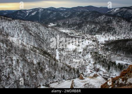 La natura si è gelata in previsione della primavera. Inverno cupo paesaggio naturale al crepuscolo. Villaggio tra le colline, vista dalla collina. Strada, fiume ghiacciato e bui Foto Stock