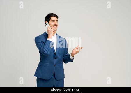 Un uomo d'affari allegro in costume blu parla sul suo telefono cellulare con un gesto della mano che indica una conversazione vivace e positiva, impostato su un semplice sfondo per concentrarsi sull'aspetto della comunicazione Foto Stock