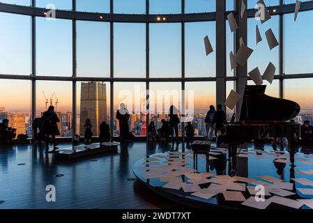 Vista panoramica attraverso una grande finestra sulle colline di Roppongi, con i visitatori che si godono la luce della sera, le sagome e il pianoforte a coda, Tokyo, Honshu, Giappone Foto Stock