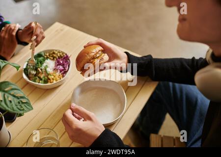 giovane uomo dal taglio corto che si gusta un pasto vegano mentre tiene in mano un hamburger con tofu vicino a una donna sfocata che mangia insalata Foto Stock