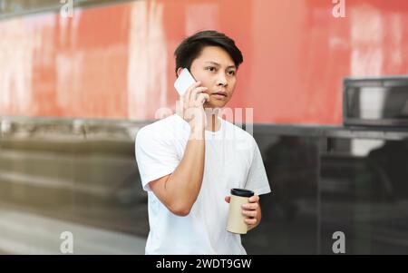 Concentrato ragazzo asiatico che parla al telefono, beve caffè Foto Stock
