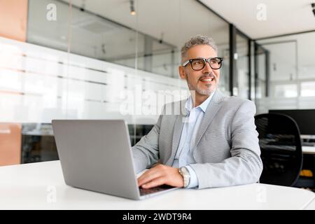 Il professionista maturo, con un sorriso gentile, ama il suo lavoro, seduto di fronte a un computer portatile in un moderno ambiente di ufficio, il che dimostra il piacere e la soddisfazione che si trovano nella cultura del lavoro flessibile di oggi Foto Stock