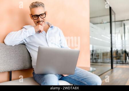 Un uomo d'affari maturo siede comodamente con un laptop sul divano in un ambiente di lavoro informale, riflettendo la tendenza moderna degli ambienti di lavoro flessibili, un equilibrio tra produttività e concetto di comfort Foto Stock