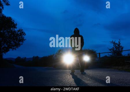 Un misterioso personaggio con cappuccio, sagomato contro i fari di un'auto parcheggiata. Su una strada di campagna solitaria e spettrale di notte. Foto Stock