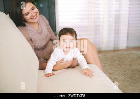 Ritratto al coperto di una giovane madre felice che gioca con un adorabile bambino, coccolandosi su un comodo divano bianco in una stanza amorevole Foto Stock