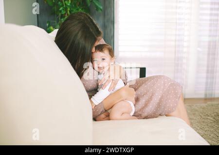 Ritratto al coperto di una giovane madre felice che gioca con un adorabile bambino, coccolandosi su un comodo divano bianco in una stanza amorevole Foto Stock