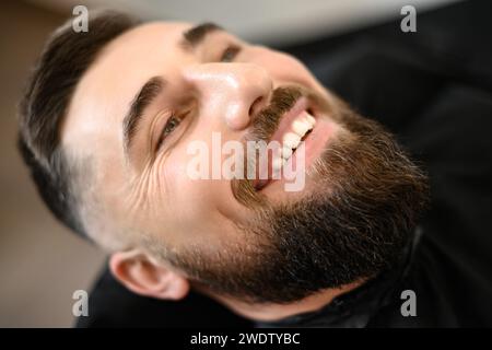 Un giovane con la barba in un mantello nero siede su una sedia in un barbiere. Il cliente sta aspettando un taglio di capelli e la rasatura della barba. Foto Stock