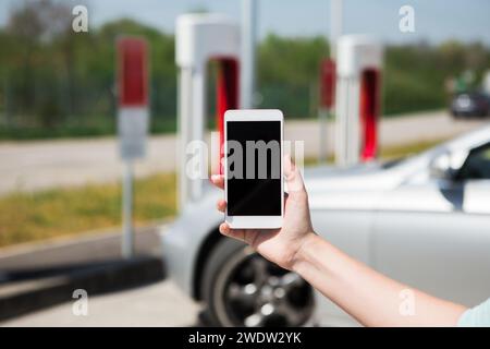 Mano con il telefono sullo sfondo della stazione di ricarica per auto elettriche. Foto Stock