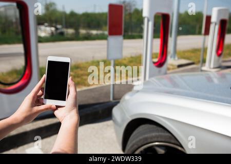 Mano con il telefono sullo sfondo della stazione di ricarica per auto elettriche. Foto Stock