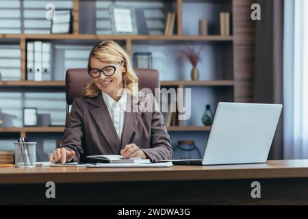 Un'elegante donna anziana in abbigliamento da lavoro sorride mentre lavora al suo computer portatile in un ambiente di ufficio domestico ben arredato, trasudando fiducia. Foto Stock