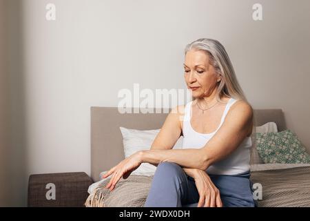 Donna anziana in casuali seduta a letto e guardando in basso. Una donna triste è seduta nella sua camera da letto. Foto Stock